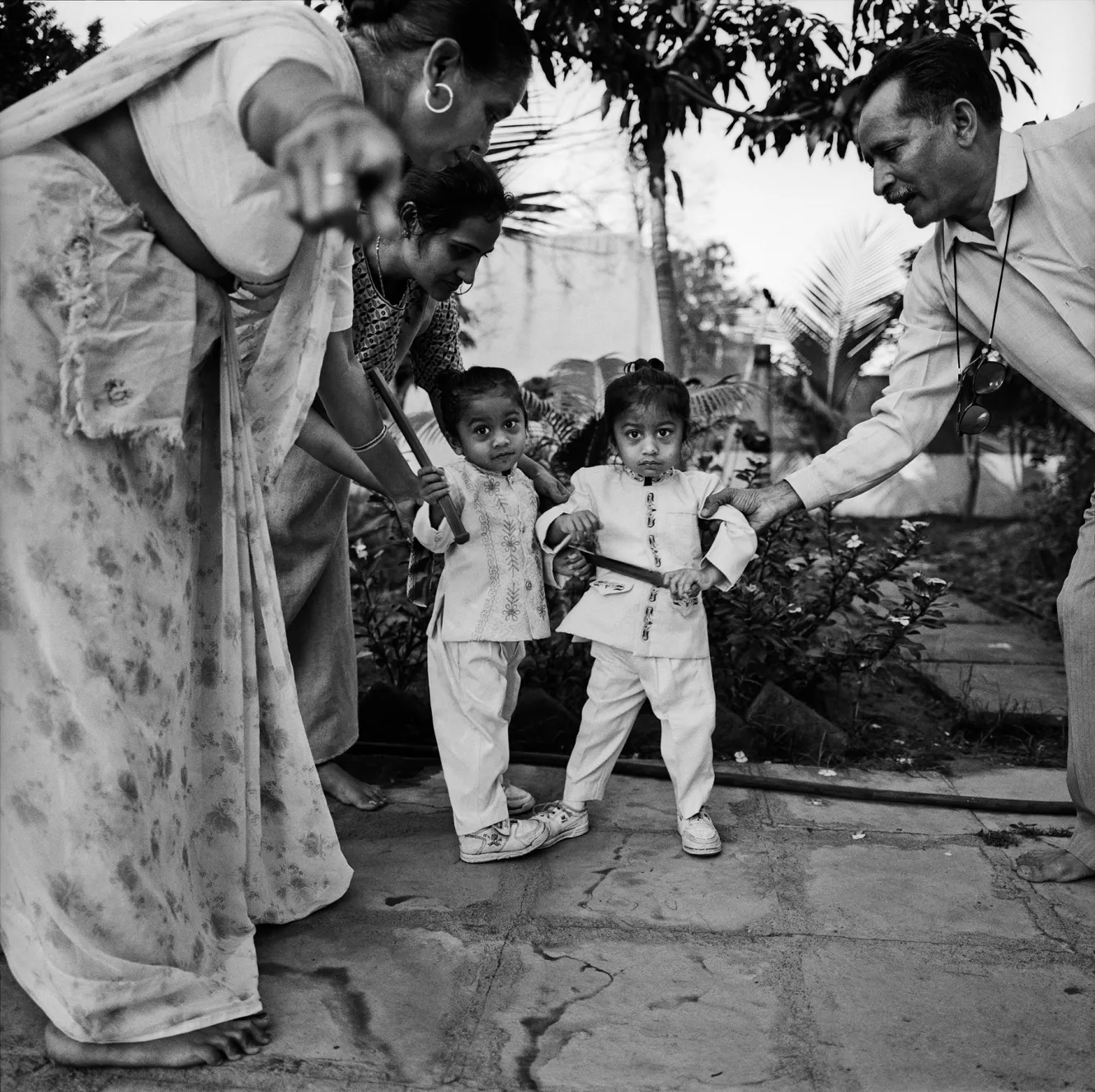 Two adults help two young children, Sanchit and Sawan, hold wooden sticks in a courtyard.