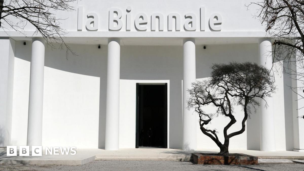 A white building bearing the words "la Biennale" with a black tree in front of it.