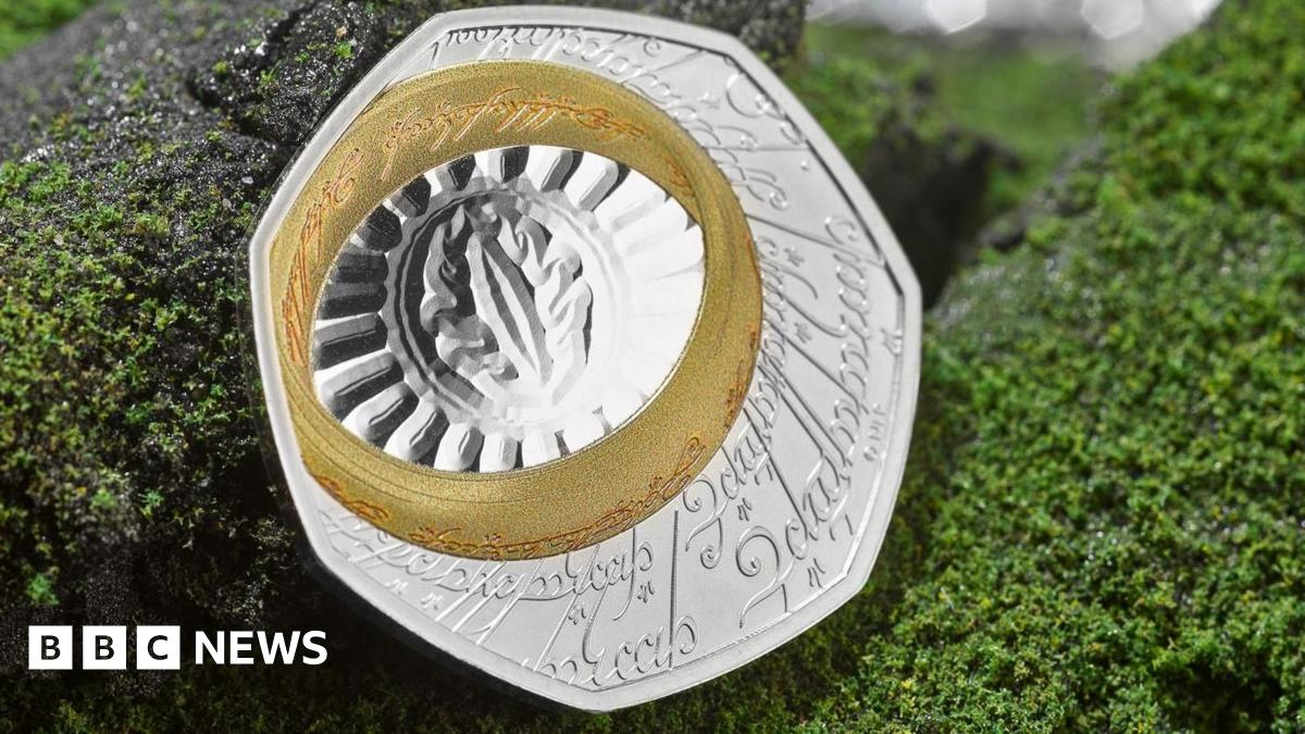 Both sides of the coin shown on a fake background of granite grey rock frosted in green lichen. The back of the coin on the left is silver with an etching in a Lord of the Rings script and language. A gold band, the one ring, is also inscribed in an Tolkien language, and sits slightly to the top left corner of the seven sided coin. it surrounds the eye of sauron, a slit etched onto the silver with an oval around it that is then surrounded by a fan of lines to the ring, making it look like the sun. the face of the coin, the right is the same as a 50p coin with a King Charles the third profile and the visible inscription fromt he top clockwise of G Rex   F D 50 pence 2026 Char with the rest of the king's name obstructed by the back of the coin.
