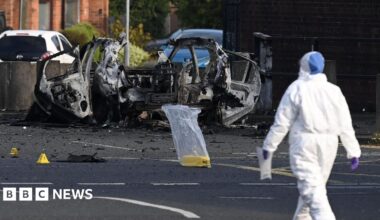A forensic investigator dressed in a white hazmat suit and purple plastic gloves gathers evidence at the scene of a car bomb attack on Dunmurry Police Station. The investigator is walking towards the wreckage of the vehicle which has been blown apart.  Three yellow evidence markers have been placed on the road, one is inside a clear plastic bag.