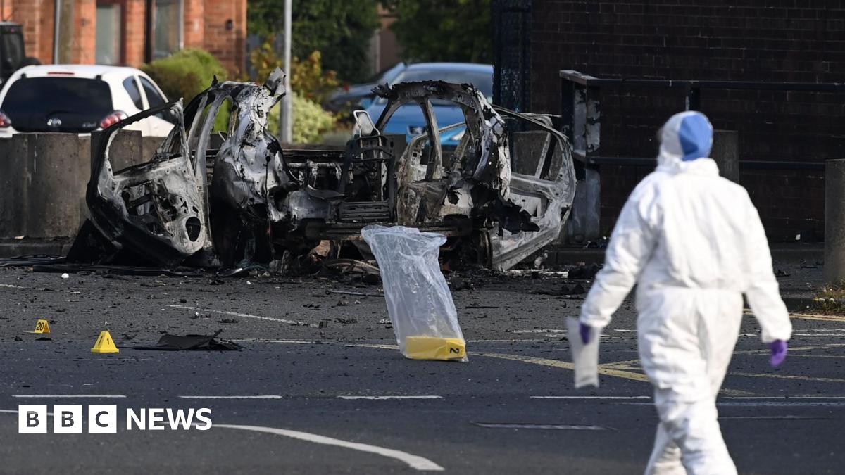 A forensic investigator dressed in a white hazmat suit and purple plastic gloves gathers evidence at the scene of a car bomb attack on Dunmurry Police Station. The investigator is walking towards the wreckage of the vehicle which has been blown apart.  Three yellow evidence markers have been placed on the road, one is inside a clear plastic bag.