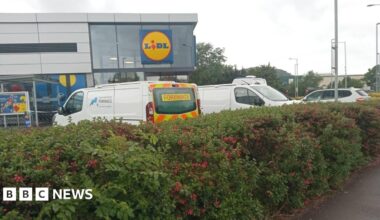 A photo of a large Lidl supermarket. Police vehicles are parked in the car park, including one marked "forensics". A police cordon is visible. A hedge surrounding the car park sits in the foreground.