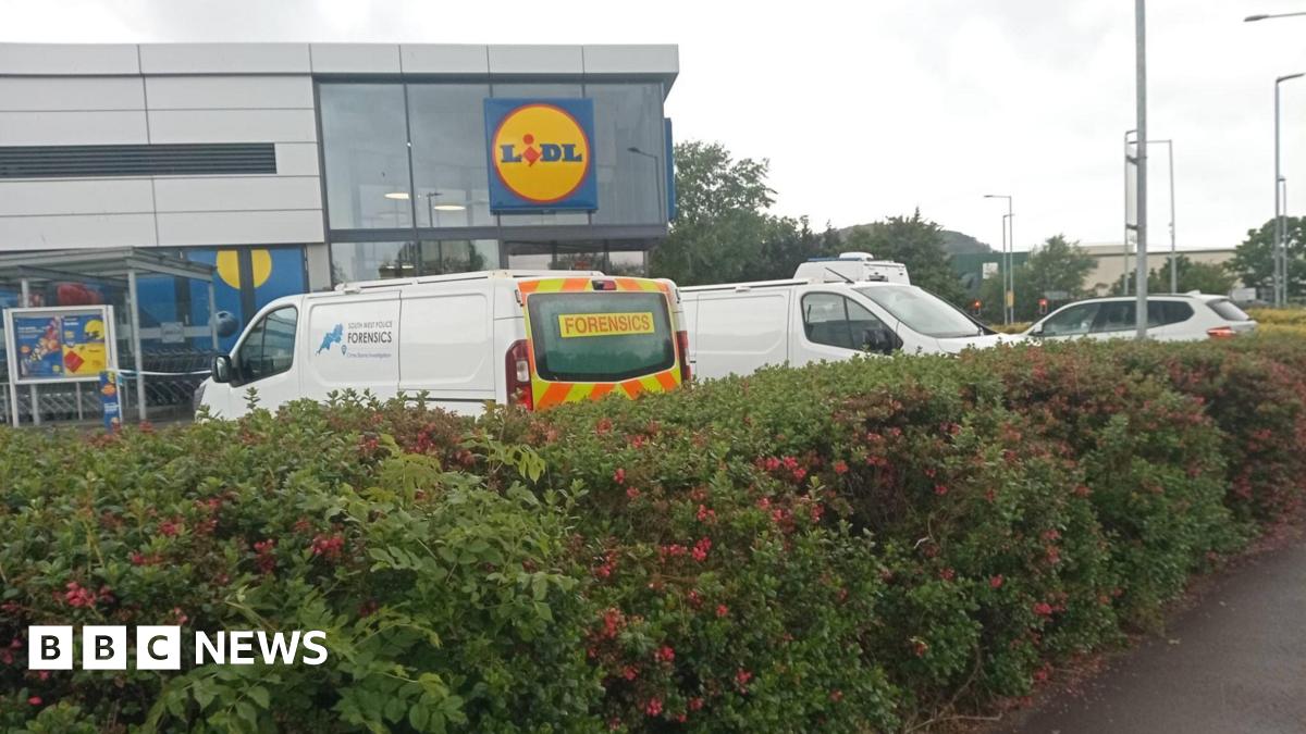 A photo of a large Lidl supermarket. Police vehicles are parked in the car park, including one marked "forensics". A police cordon is visible. A hedge surrounding the car park sits in the foreground.
