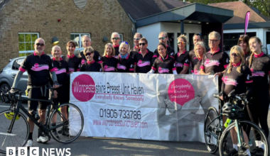 About 20 cyclists wearing mainly black outfits stand with bikes and hold up a large mainly white banner including the words Worcestershire Breast Unit Haven. Everybody knows somebody are among other words printed on the banner.