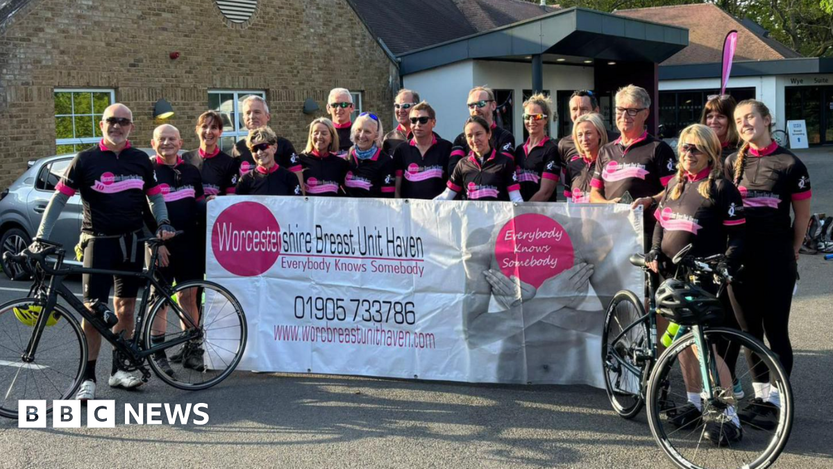 About 20 cyclists wearing mainly black outfits stand with bikes and hold up a large mainly white banner including the words Worcestershire Breast Unit Haven. Everybody knows somebody are among other words printed on the banner.
