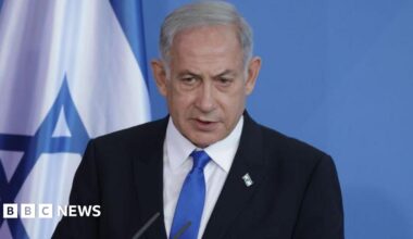 A shot of Netanyahu from the shoulders up, wearing a dark suit with a white shirt and a light blue tie. He has a small pin of the Israel flag on his lapel. Behind him is a plain blue background with part of the Israel flag to the left.