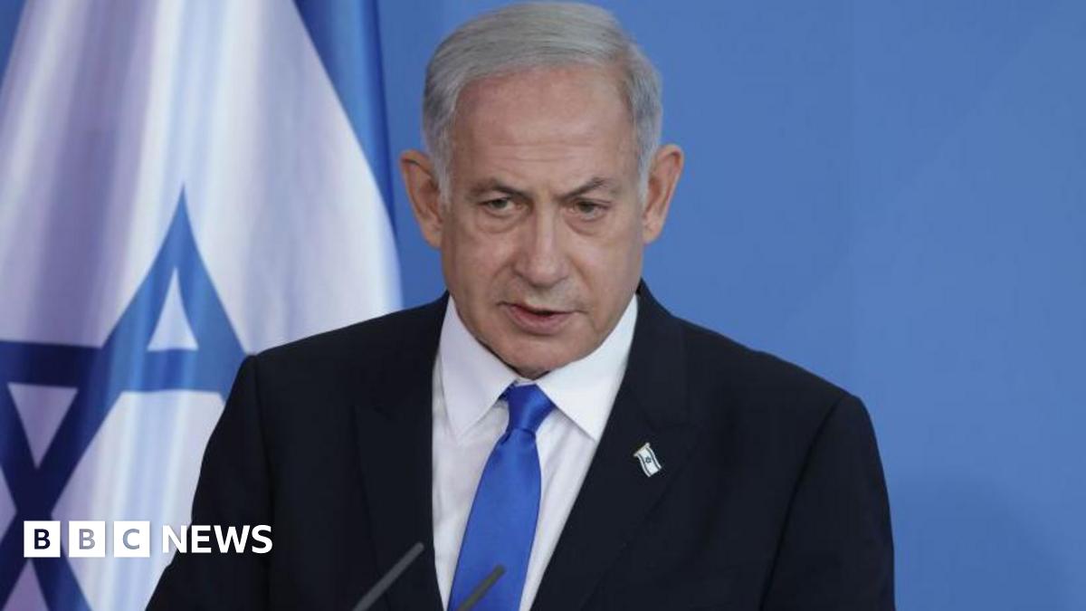 A shot of Netanyahu from the shoulders up, wearing a dark suit with a white shirt and a light blue tie. He has a small pin of the Israel flag on his lapel. Behind him is a plain blue background with part of the Israel flag to the left.