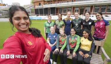 Afra Rose Mohamed has long dark hair and is wearing a red cricket top. This picture is a selfie with her in the foreground and 10 other girls smiling in the background. They are on the pitch at Lord's.