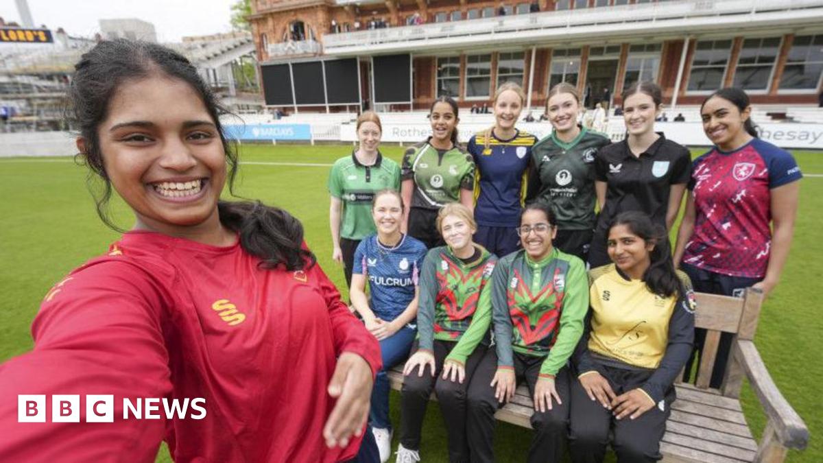 Afra Rose Mohamed has long dark hair and is wearing a red cricket top. This picture is a selfie with her in the foreground and 10 other girls smiling in the background. They are on the pitch at Lord's.