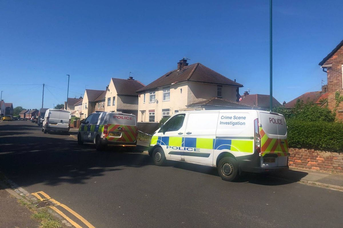 A police van is parked on a residential street, with a house visible in the background. The sky is clear, and the road is lined with other buildings and greenery.