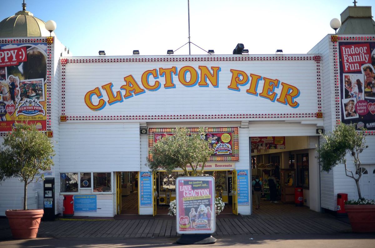 The entrance of a vibrant amusement park, prominently displaying the name "Clacton Pier" in bold yellow letters against a white backdrop. The façade is decorated with colorful posters and illuminated by overhead lights. In front of the entrance, there is a small stand with a sign and two potted trees adding a touch of greenery to the scene.