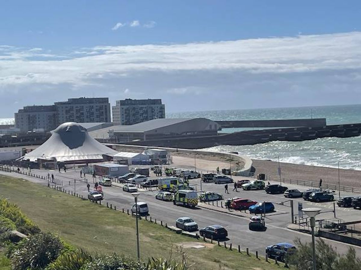 An aerial view of a coastal city with modern architecture, a prominent curved building, and a busy road leading to a car park with numerous vehicles parked along the seashore.