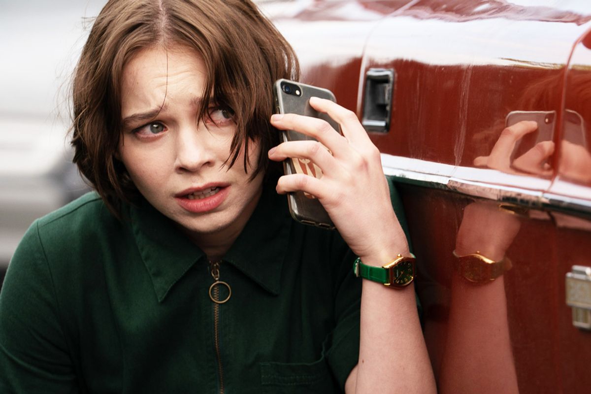 Teenage girl looking worried crouches by red car with smartphone held up to her ear