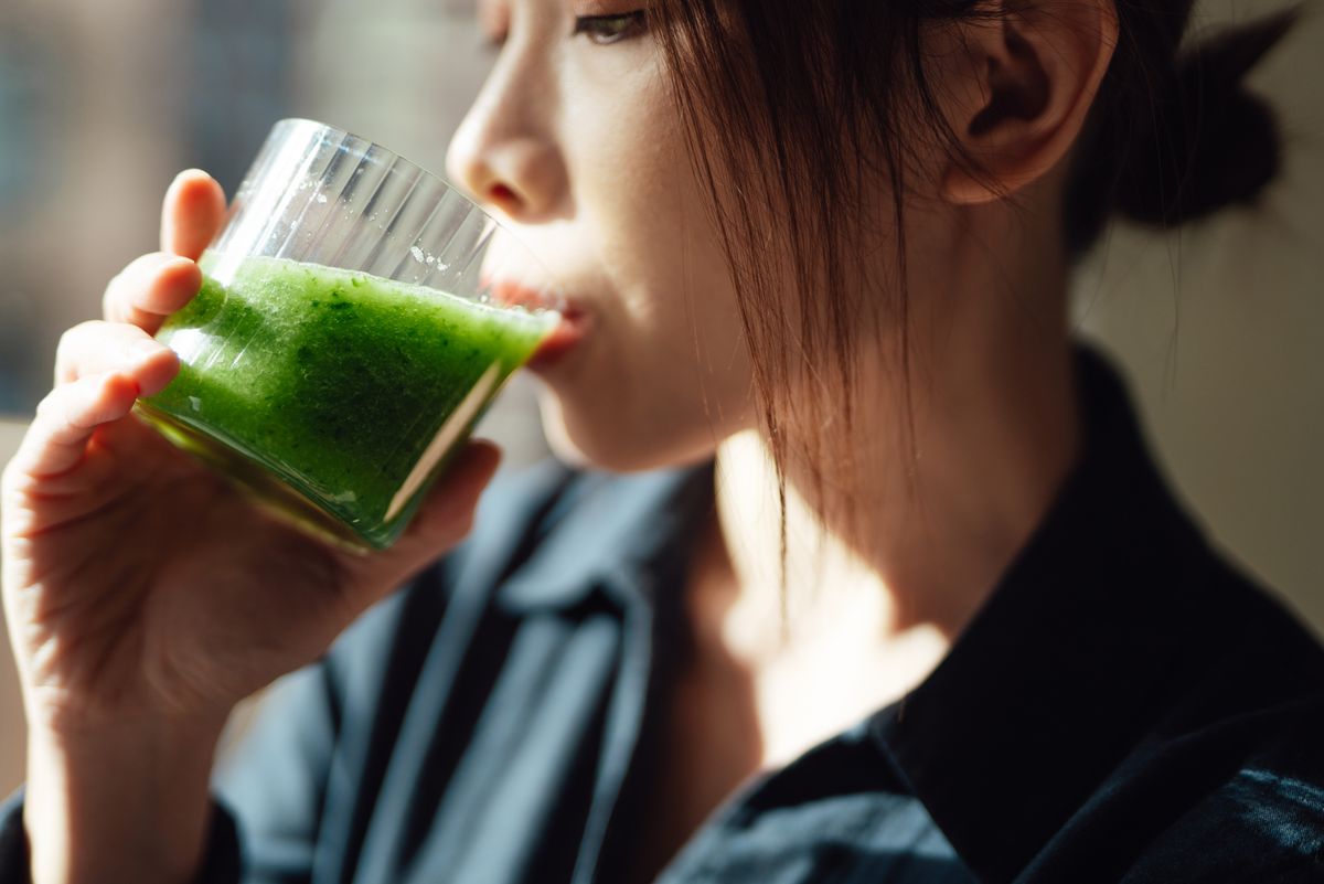 Young Asian woman drinking a glass of fresh green fruit and vegetable smoothie with natural sunlight. Healthy superfood. Detox diet.