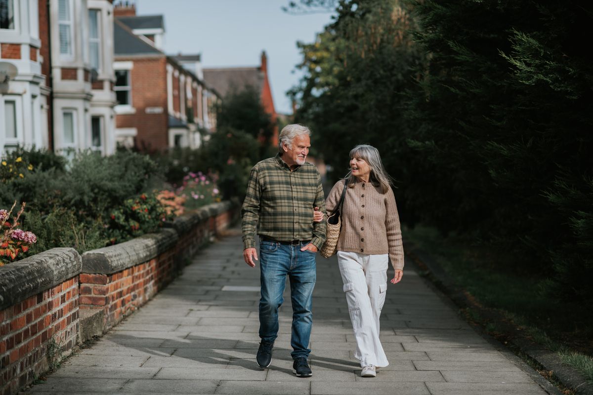 A mature man and woman stroll down a footpath beside a row of terrace houses in a beautiful residential district. They cast a shadow on the street as they have a lighthearted discussion.