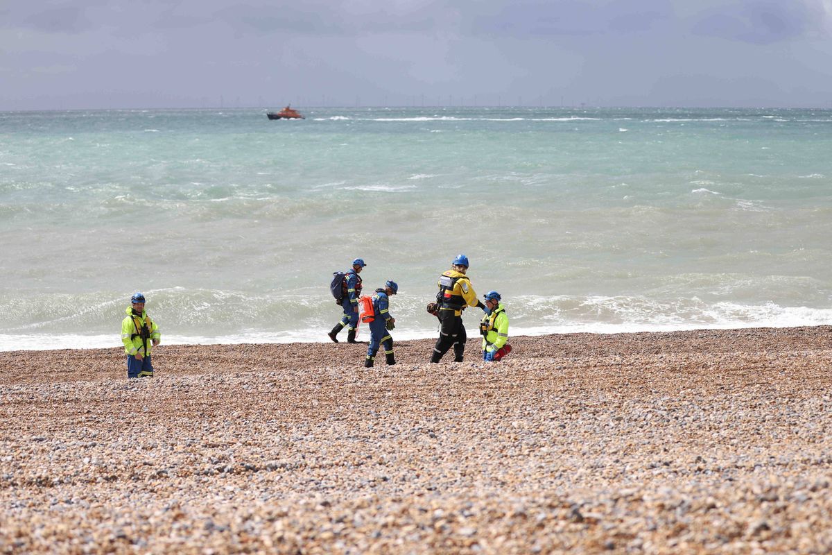A group of individuals, dressed in high-visibility safety gear, are gathered on a pebbled beach, with the ocean visible in the background. The individuals appear to be engaged in an activity, possibly a search or rescue operation. A small boat can be seen on the water in the distance, suggesting the presence of a coordinated effort in the area.