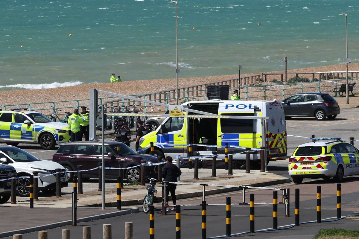 An aerial view of a beach area where several police vehicles are parked along the roadside, with a beach and ocean visible in the background.