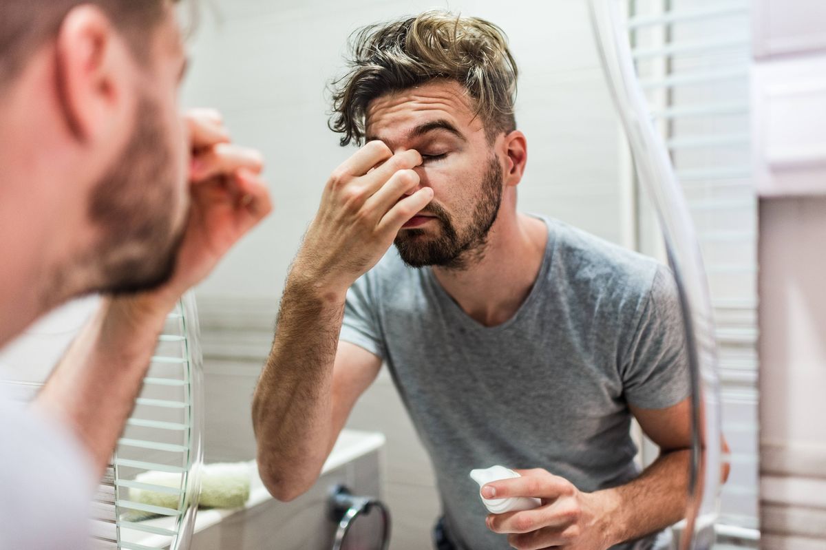 Shot of a young man standing in front of the bathroom mirror looking exhausted.
