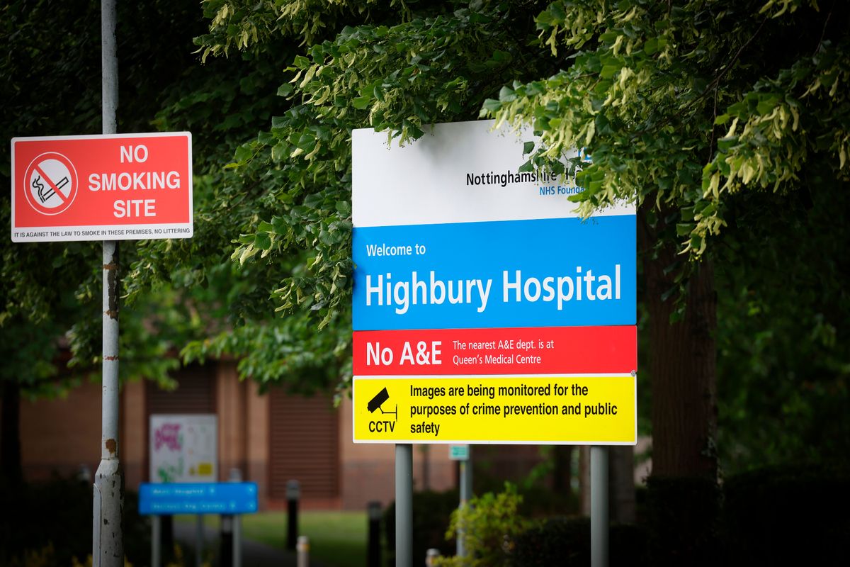 NHS sign with white, blue, red and yellow horizontal stripes and text on, at entrance to hospital, with "no smoking site" sign on left, with thick tree covering behind and buildings visible blurred in background