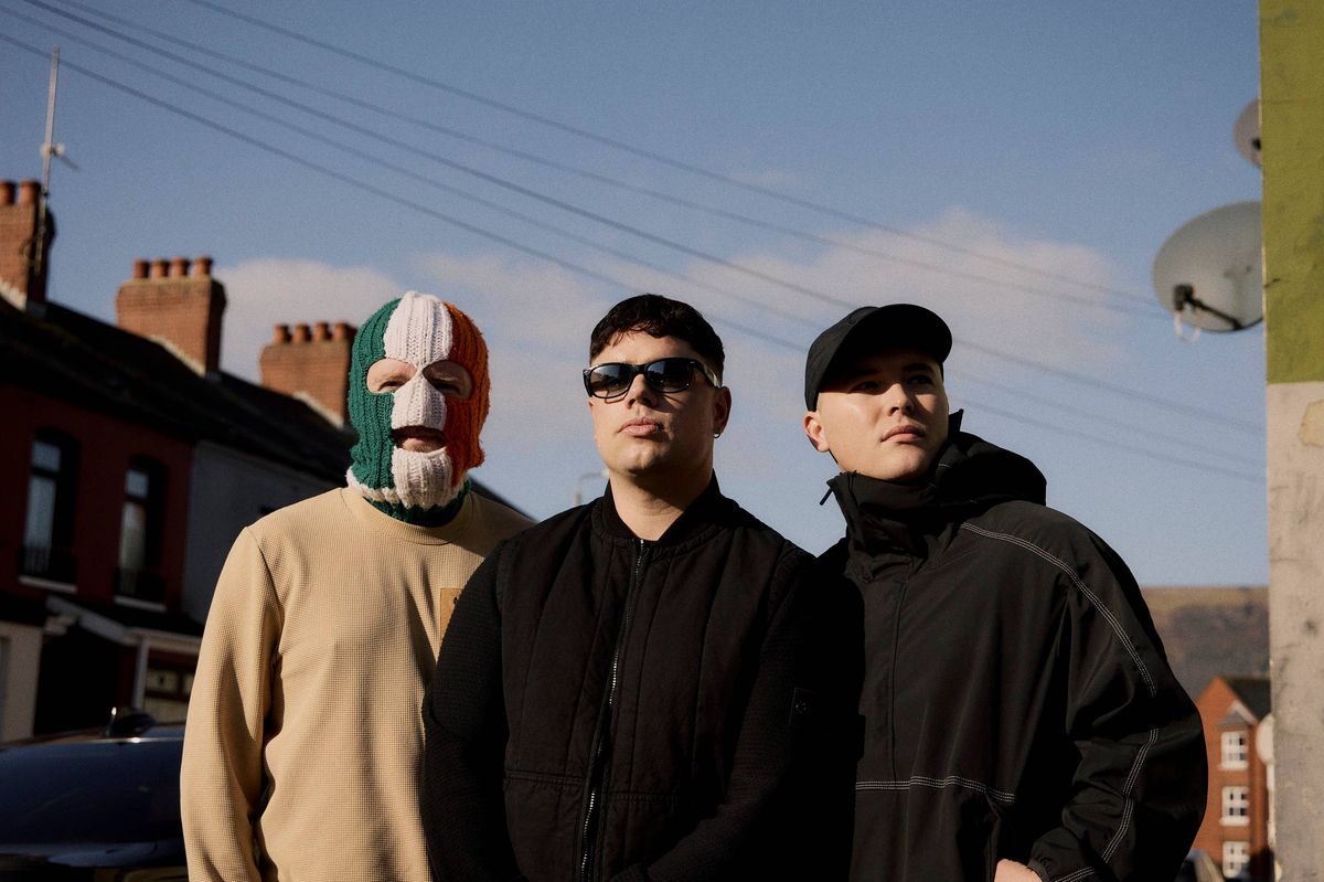 Three individuals, dressed in varying outerwear, stand in close proximity in an urban setting. One individual dons a mask in the colors of the Irish flag, while the other two individuals wear sunglasses and caps. Behind them, houses and electric transmission lines are visible against a partly cloudy sky.