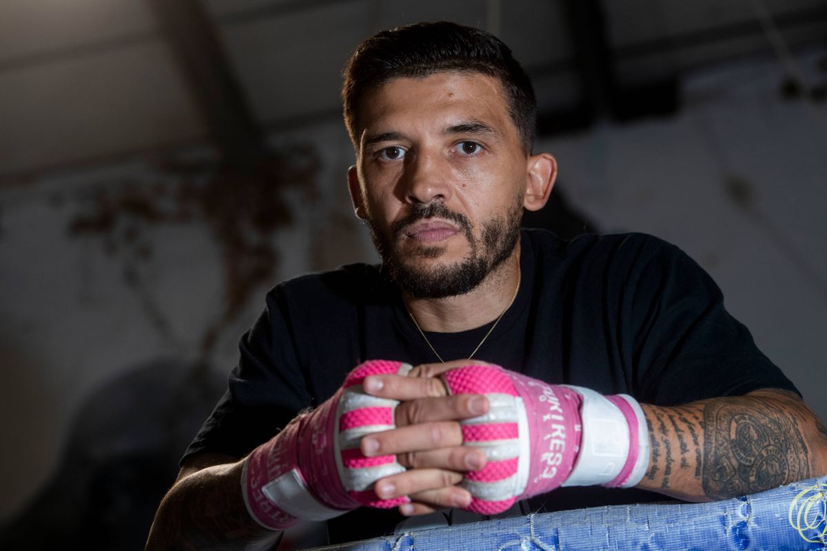 An individual with a serious expression, wearing a black t-shirt and pink boxing gloves, is seated in a dimly lit room, leaning on a blue surface.