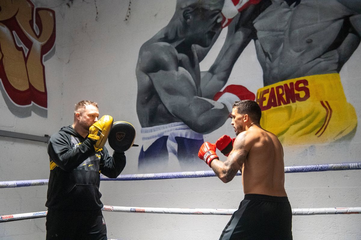 Two individuals are engaged in a boxing training session inside a gym. One person, wearing a black jacket and gloves, appears to be providing instructions or coaching. The other person, shirtless and wearing black shorts, is wearing red gloves and is likely practicing punches. A large mural of a muscular boxer is prominently displayed on the wall behind them, adding to the thematic setting of the training environment.