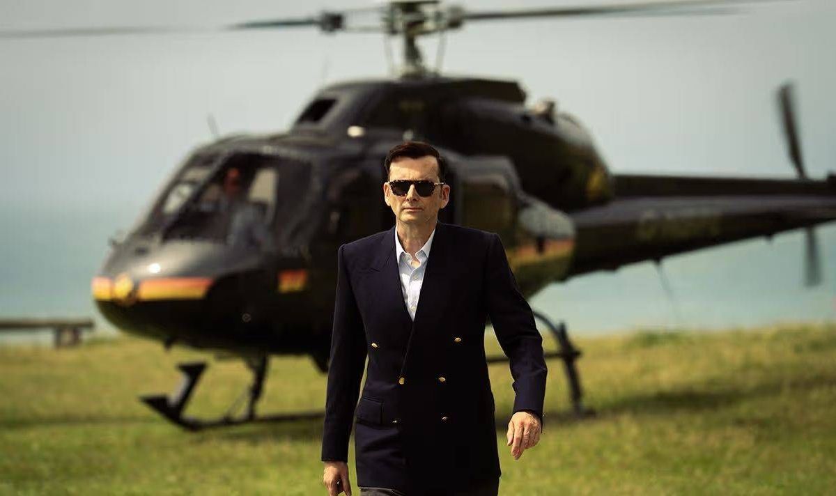 An individual dressed in a formal black suit is walking towards a black helicopter parked on a grassy field, under a clear sky.