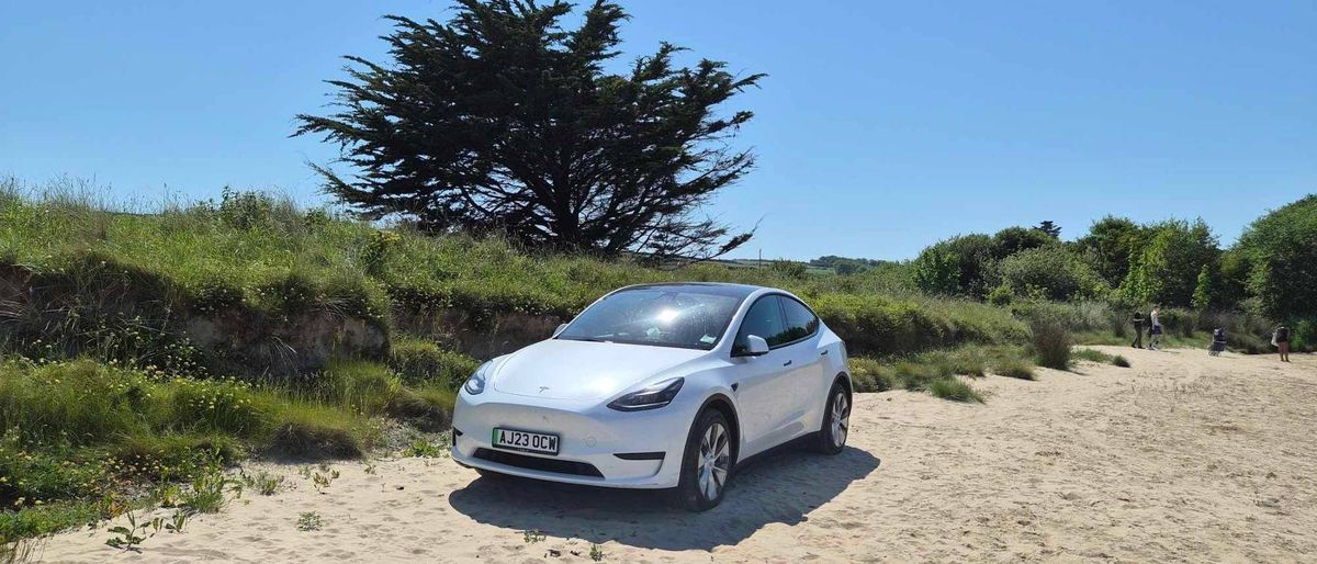 A white, modern, four-door electric vehicle is parked on a dirt road with a backdrop of a grassy hill and a single prominent tree under a clear blue sky.