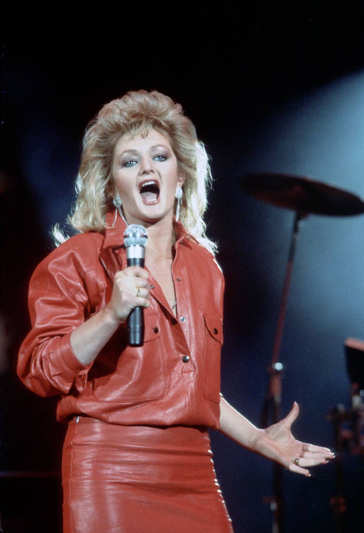 An individual with blonde hair, dressed in a red jacket, is performing on stage, holding a microphone and singing passionately. A cymbal is visible in the background.