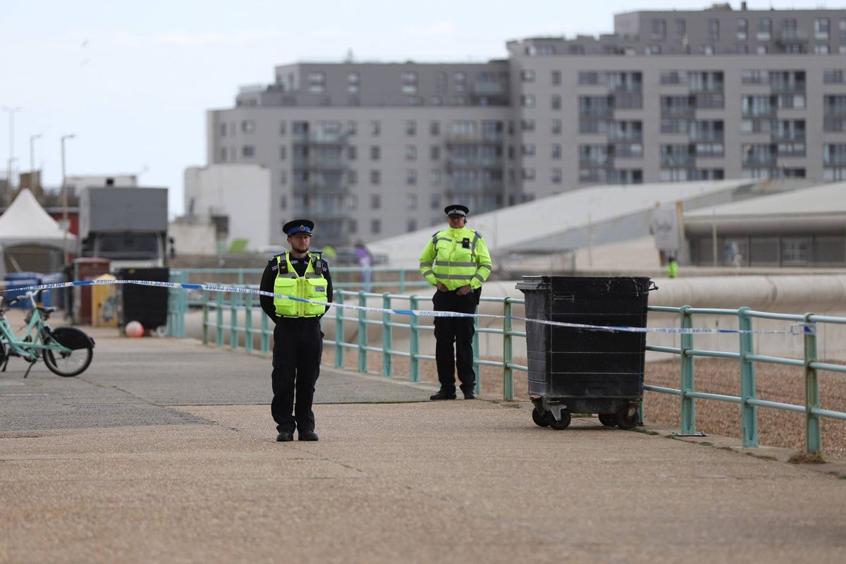 Two individuals in high-visibility vests stand guard on a paved area, with a trash receptacle and a bicycle in the background. A barricade and a large building are visible in the distance, suggesting a controlled or secure environment.