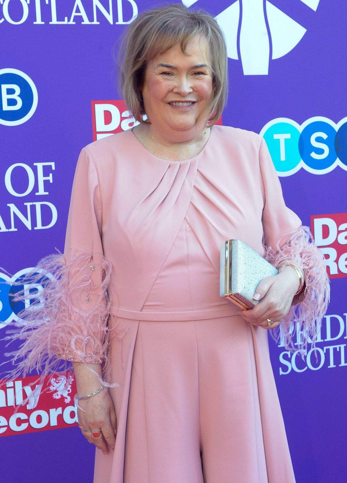 An adult woman, smiling and standing, is captured in front of a vibrant purple backdrop adorned with various logos and text. The woman is dressed in a pink blouse and her expression exudes happiness and contentment.