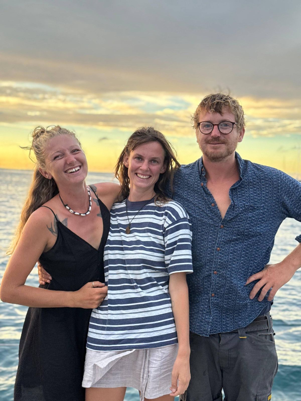 Three individuals are standing close together on a beach, with a serene ocean and a colorful sunset in the background. The individual on the left is wearing a sleeveless black dress and has long hair. The middle person is dressed in a white and blue striped shirt and white shorts, with their hair tied back. The person on the right is wearing a blue shirt and has a mustache. They appear to be posing for a photograph.