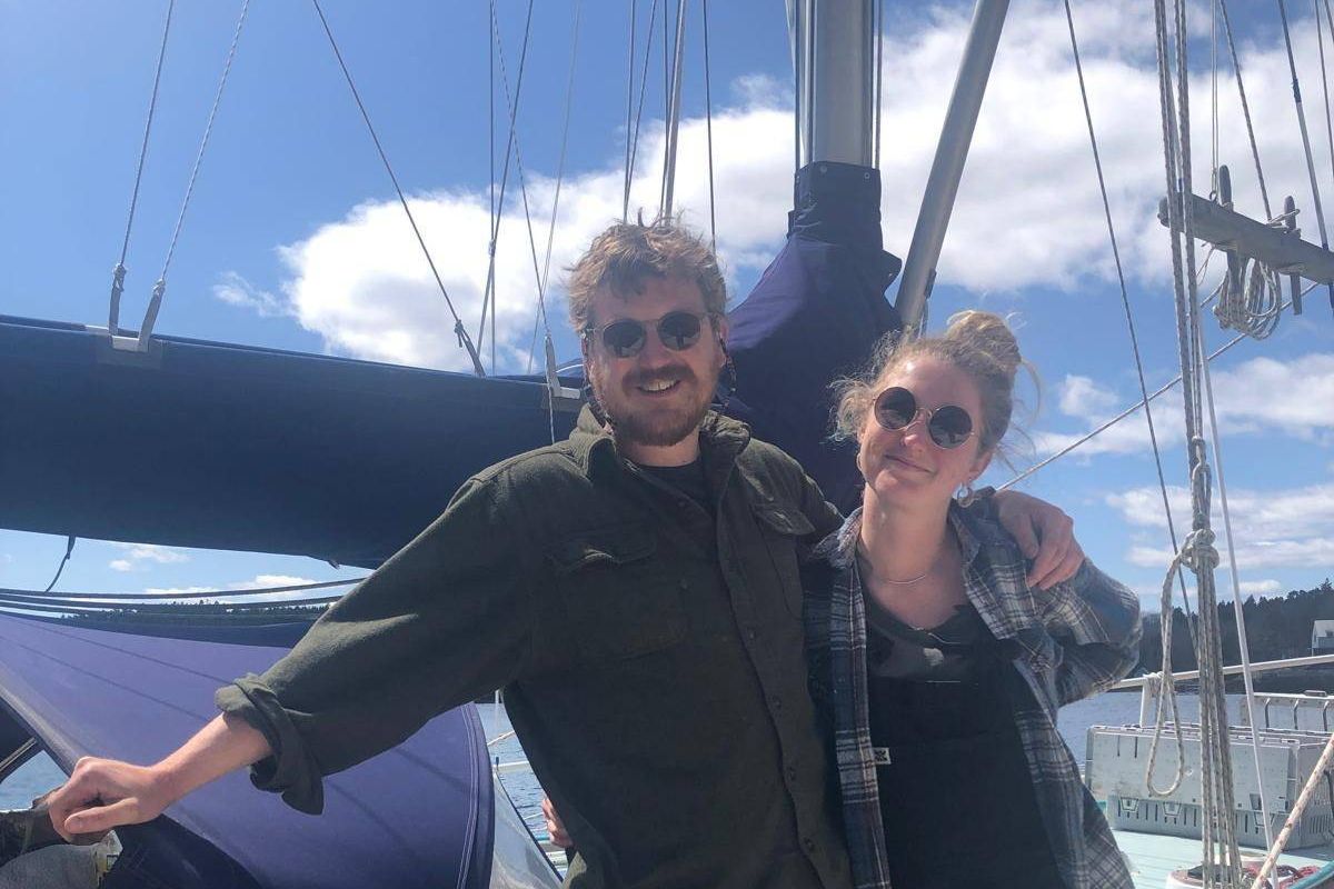 Two individuals, likely a couple, are standing on the deck of a sailboat, with one individual's arm around the other's shoulder, under a clear sky with some clouds.