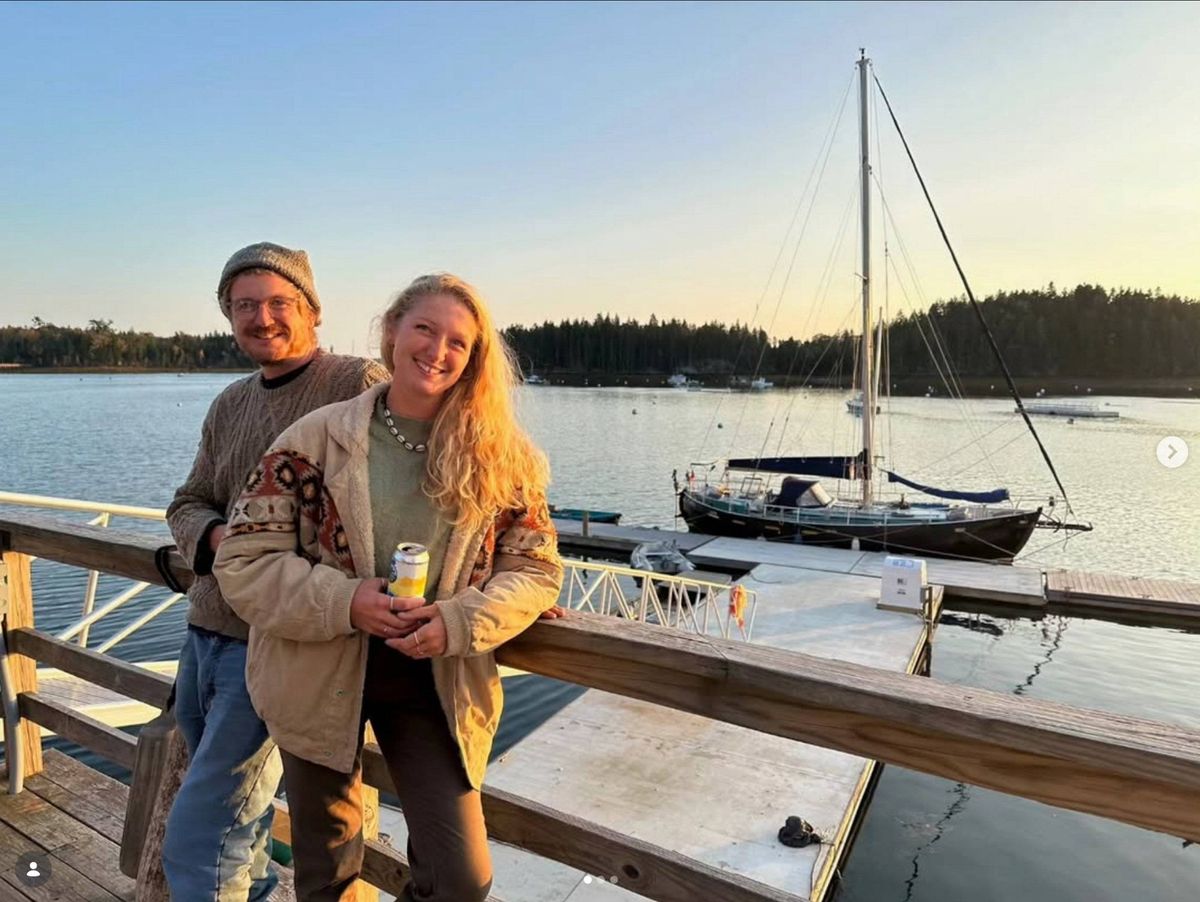 Two individuals, one male and one female, are standing together on a wooden pier by the water, with a sailboat docked nearby. The scene captures a moment of leisure against a backdrop of a serene sunset.