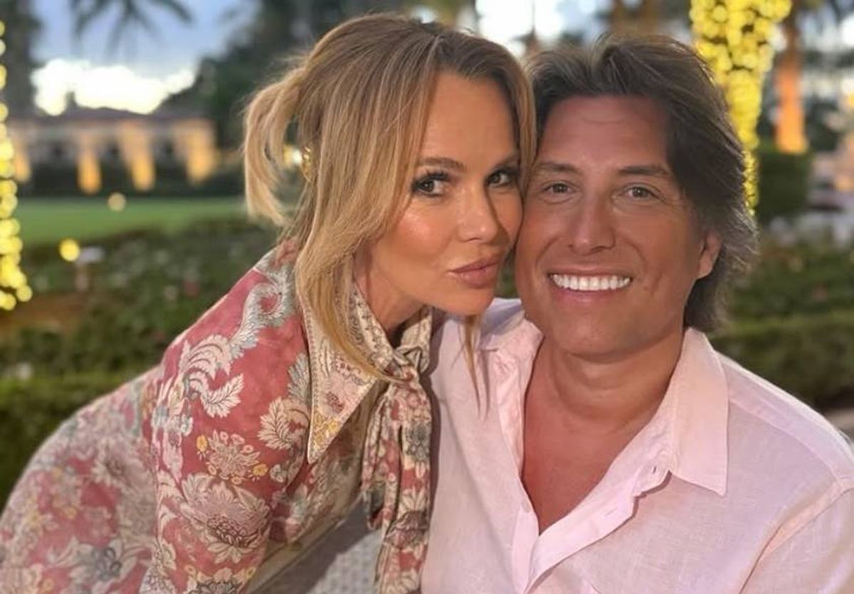 Two individuals, possibly a couple, are posing for a photograph in an outdoor setting with a backdrop of greenery and illuminated architectural features. The person on the left, with blonde hair, is leaning in close to the person on the right, who is smiling widely.