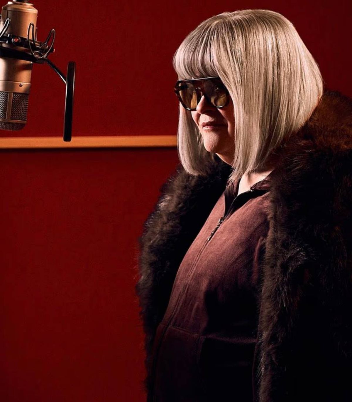 An individual with short, straight, blonde hair wearing sunglasses and a fur collared coat standing in front of a red wall.