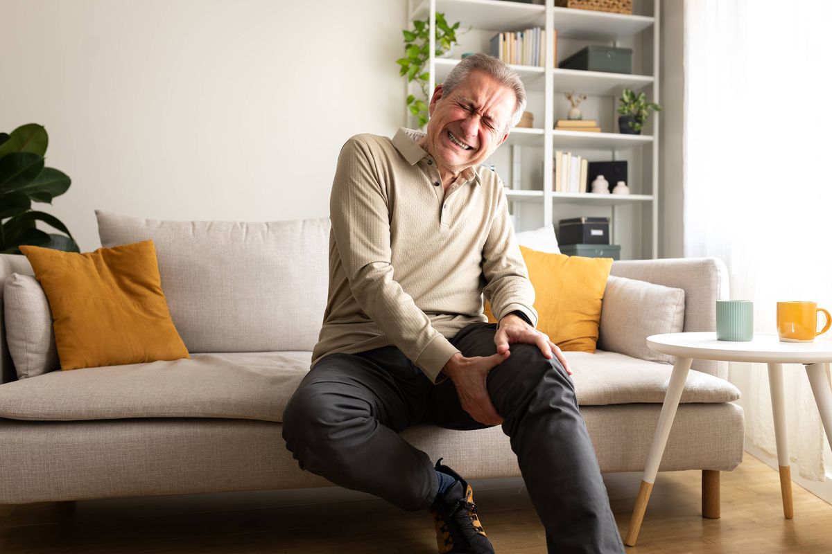 Senior man suffering from knee pain while sitting on his sofa at home, holding his leg with pained expression