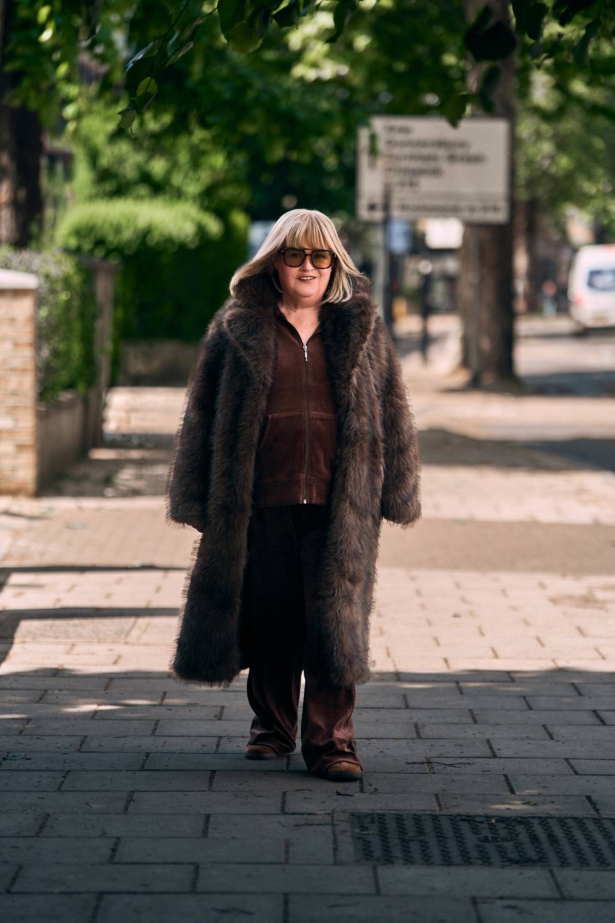 An adult female wearing a fur coat and sunglasses is walking down a city street, with a slight smile on her face.