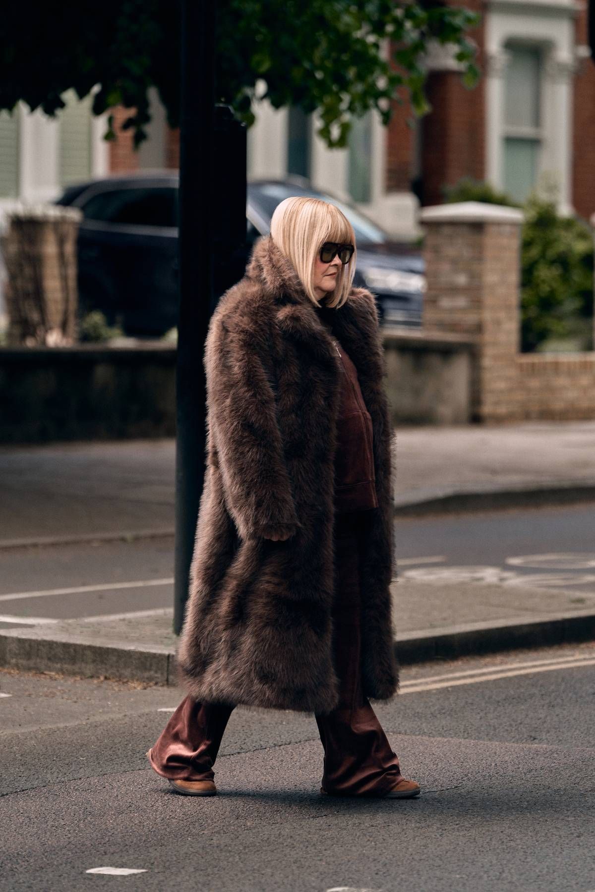 An individual, wrapped in a fur coat, is strolling along a sidewalk adjacent to a row of townhouses. The scene includes a tree and a parked vehicle in the background.