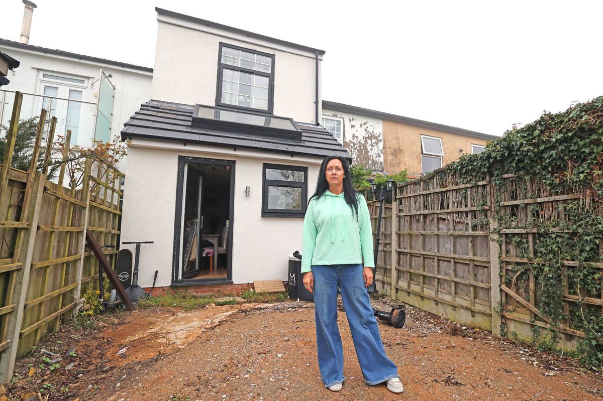An individual, dressed in a light green long-sleeved top and flared pants, is standing in front of a residential house. The house is white with a gray roof, and solar panels are installed on the roof. The individual is standing on a dirt path, with a wooden fence and greenery in the background.