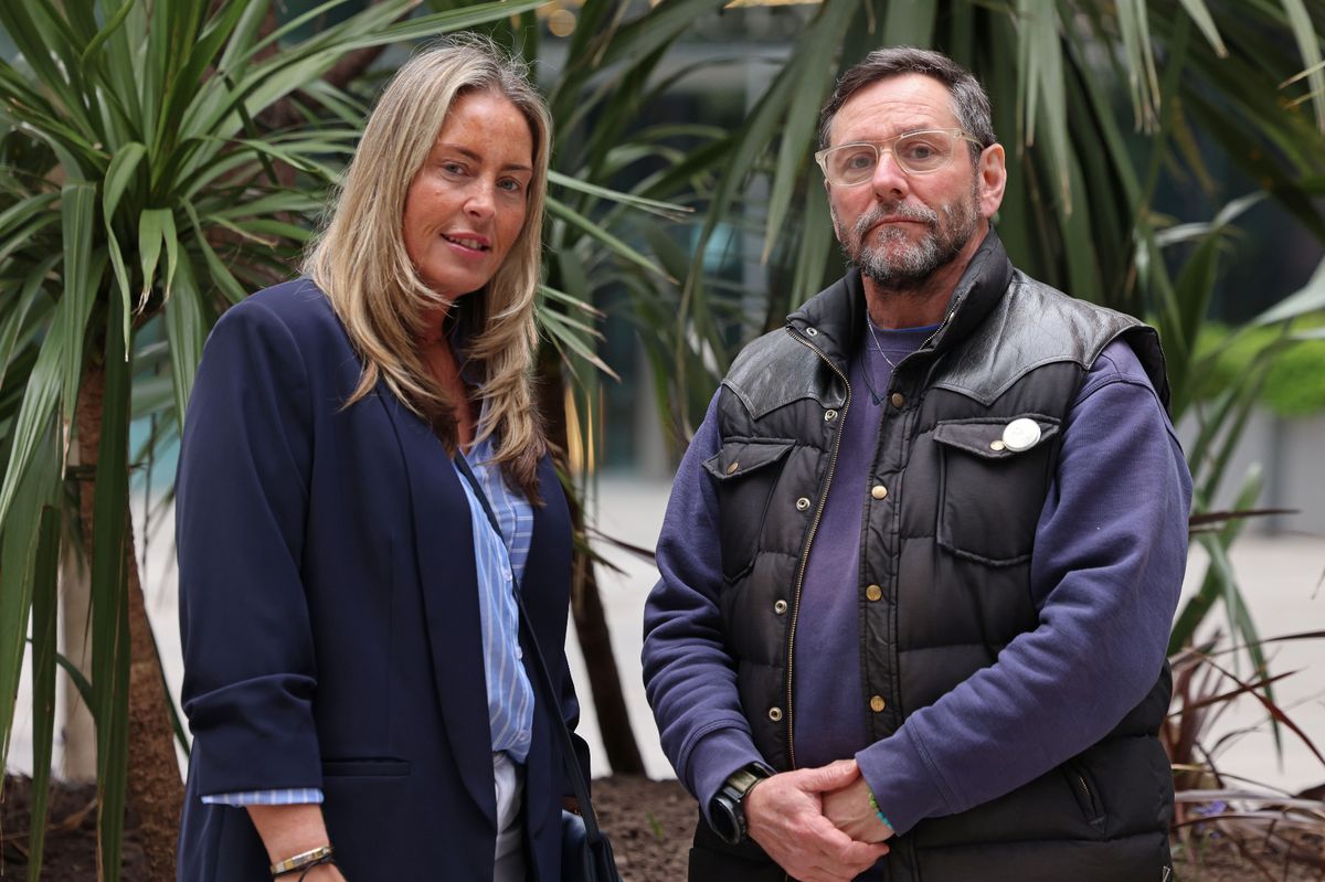 A man and woman stood against a background of foliage, they have serous faces.