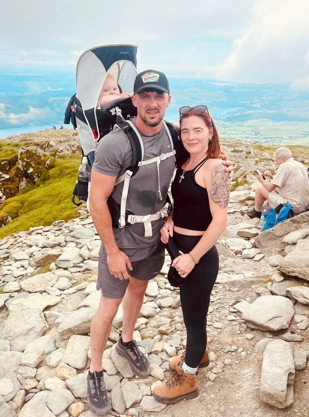 An image of a couple standing on a rocky hill, with the male individual wearing a cap and carrying a backpack. The female individual is wearing a black sleeveless top. They are smiling and posing for a photograph, with a vast landscape and sea visible in the background.