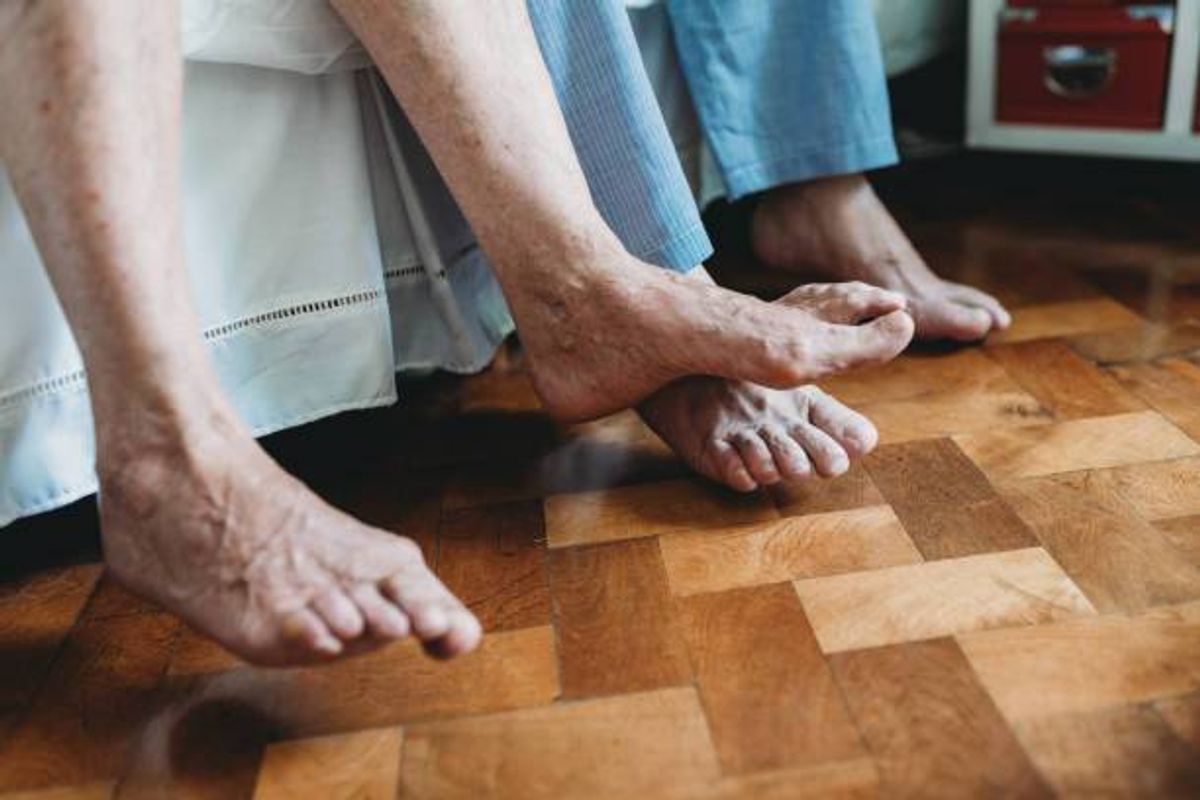 Individuals are seated on a floor, their legs and feet prominently displayed. The flooring is composed of wooden planks, and a piece of furniture is partially visible in the background.