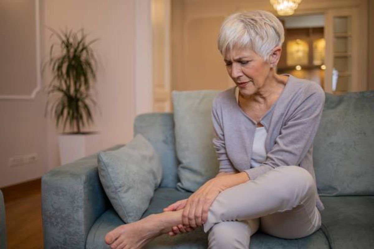 An older individual is seated on a blue couch, facing forward, with their left leg raised and resting on their right thigh, suggesting a possible injury. They appear to be in a well-lit living room, with a potted plant and a lamp visible in the background.