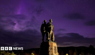The aurora borealis glows purple behind the Commando Memorial at Spean Bridge. Two of the figures on the memorial are visible. The figures are portrayed wearing military fatigues.