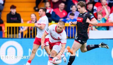 Hull KR stand-off Mikey Lewis is about to touch the ball down with his left hand for a try as a Wigan player jogs behind him