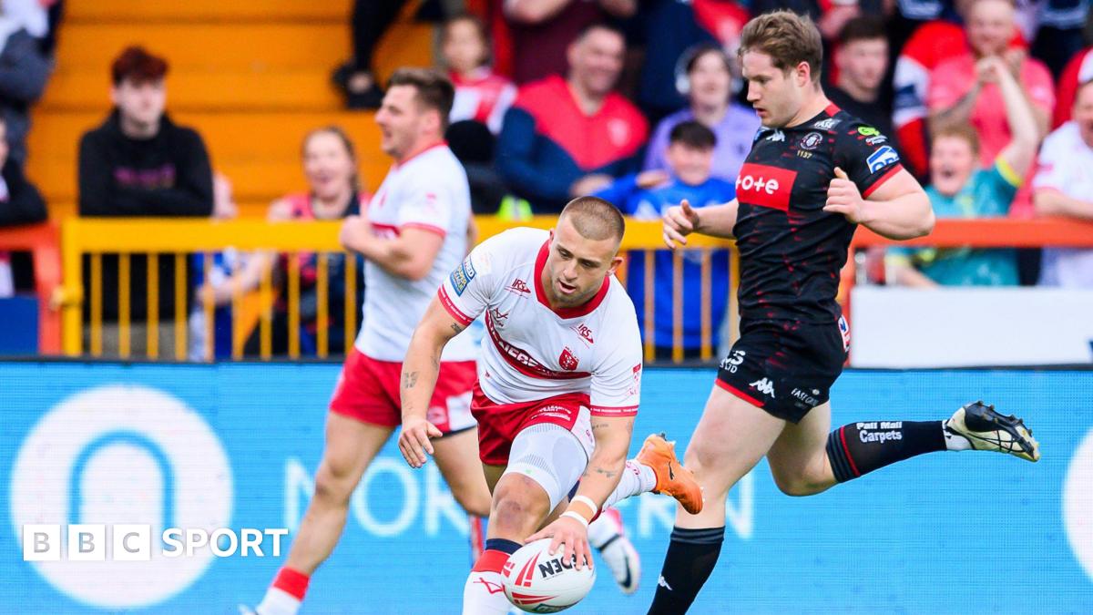 Hull KR stand-off Mikey Lewis is about to touch the ball down with his left hand for a try as a Wigan player jogs behind him