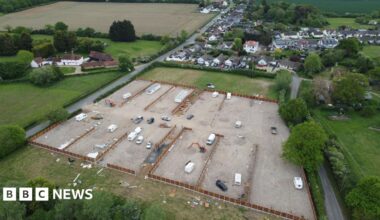 A drone image of the developed site. It is a field that has been largely concreted over and has several caravans, both static and mobile, on it. Fencing is being installed around the perimeter of the site, which is flanked by residential housing and other fields.