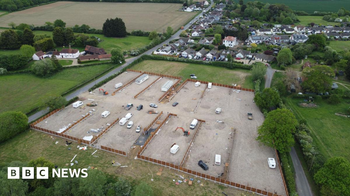 A drone image of the developed site. It is a field that has been largely concreted over and has several caravans, both static and mobile, on it. Fencing is being installed around the perimeter of the site, which is flanked by residential housing and other fields.