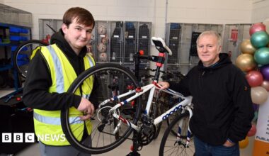 Two men standing in a bicycle repair shop. A white road bike is being held on a repair stand.